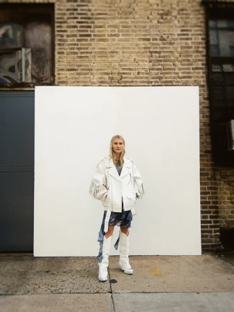 Woman in white fringed leather jacket and white cowboy boots standing against white backdrop with brick wall