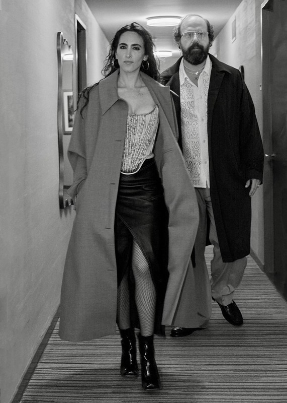 Two people in a hallway: a woman in a corset and skirt, and a man in a coat and glasses, walking toward the camera in a hotel-like hallway.