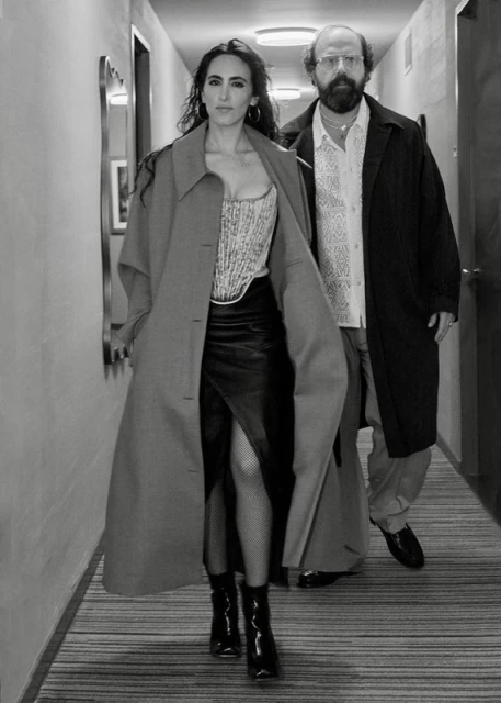 Two people in a hallway: a woman in a corset and skirt, and a man in a coat and glasses, walking toward the camera in a hotel-like hallway.
