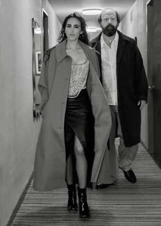 Two people in a hallway: a woman in a corset and skirt, and a man in a coat and glasses, walking toward the camera in a hotel-like hallway.