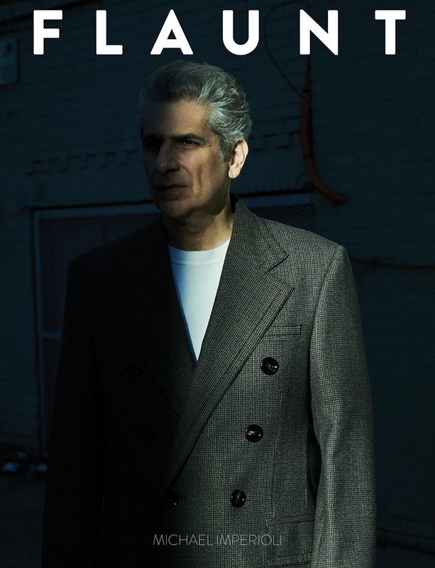 Michael Imperioli poses in front of a blue wall wearing a grey suit, white shirt and a serious expression for Flaunt magazine.