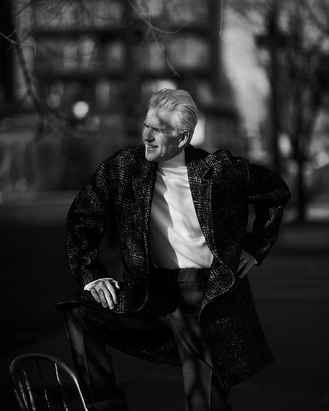 An older adult male poses in a dark coat and pants outdoors. He leans on a chair with a building visible in the background.