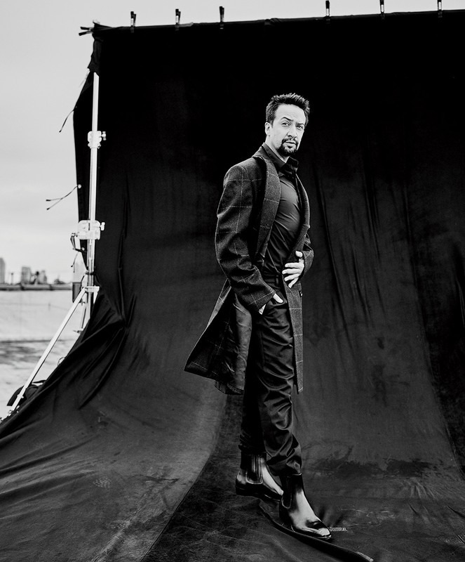 Black and white portrait of a man standing on a black backdrop wearing a suit, coat and black boots while looking at the camera.