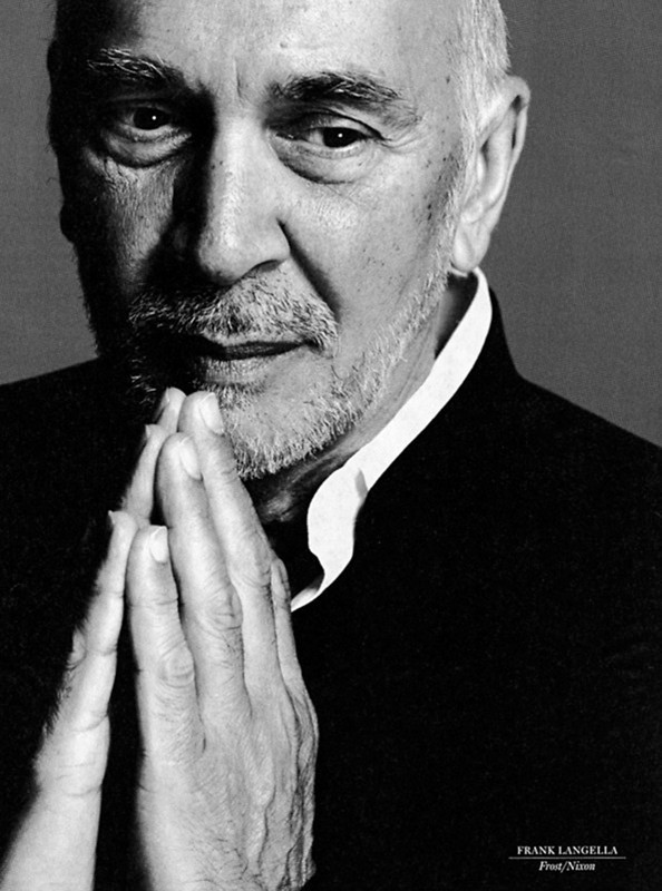 Black and white portrait of mature man with beard in suit with hands together in prayer or thoughtful pose, looking at camera.