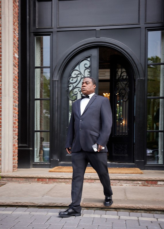 African American man in suit walks from brick mansion, holding phone, illuminated by natural light on paved stone steps.