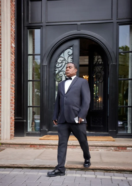 African American man in suit walks from brick mansion, holding phone, illuminated by natural light on paved stone steps.