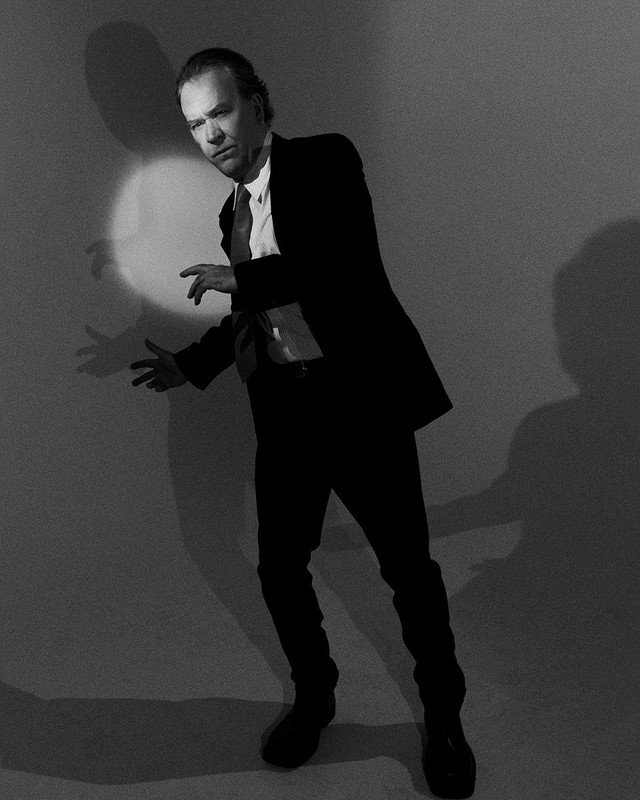 Monochrome portrait of a man in a suit gesturing on a light grey background with a spotlight and shadow, fashion or editorial style.