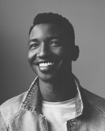 Portrait of a smiling young Black man wearing a denim jacket, shot in black and white.