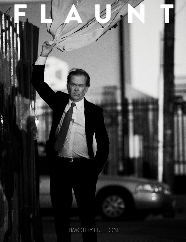 Black and white portrait of Timothy Hutton in a suit, holding a translucent object above his head, with urban background. FLAUNT magazine.