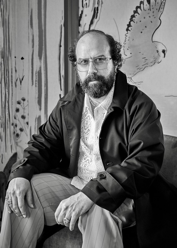 Monochrome portrait of a man with a beard, wearing glasses and a dark coat, sitting in front of a painting of a bird.