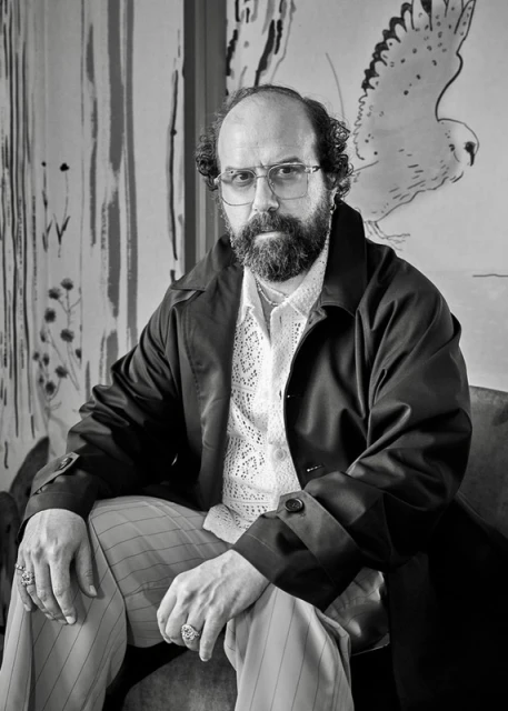 Monochrome portrait of a man with a beard, wearing glasses and a dark coat, sitting in front of a painting of a bird.