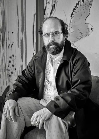 Monochrome portrait of a man with a beard, wearing glasses and a dark coat, sitting in front of a painting of a bird.