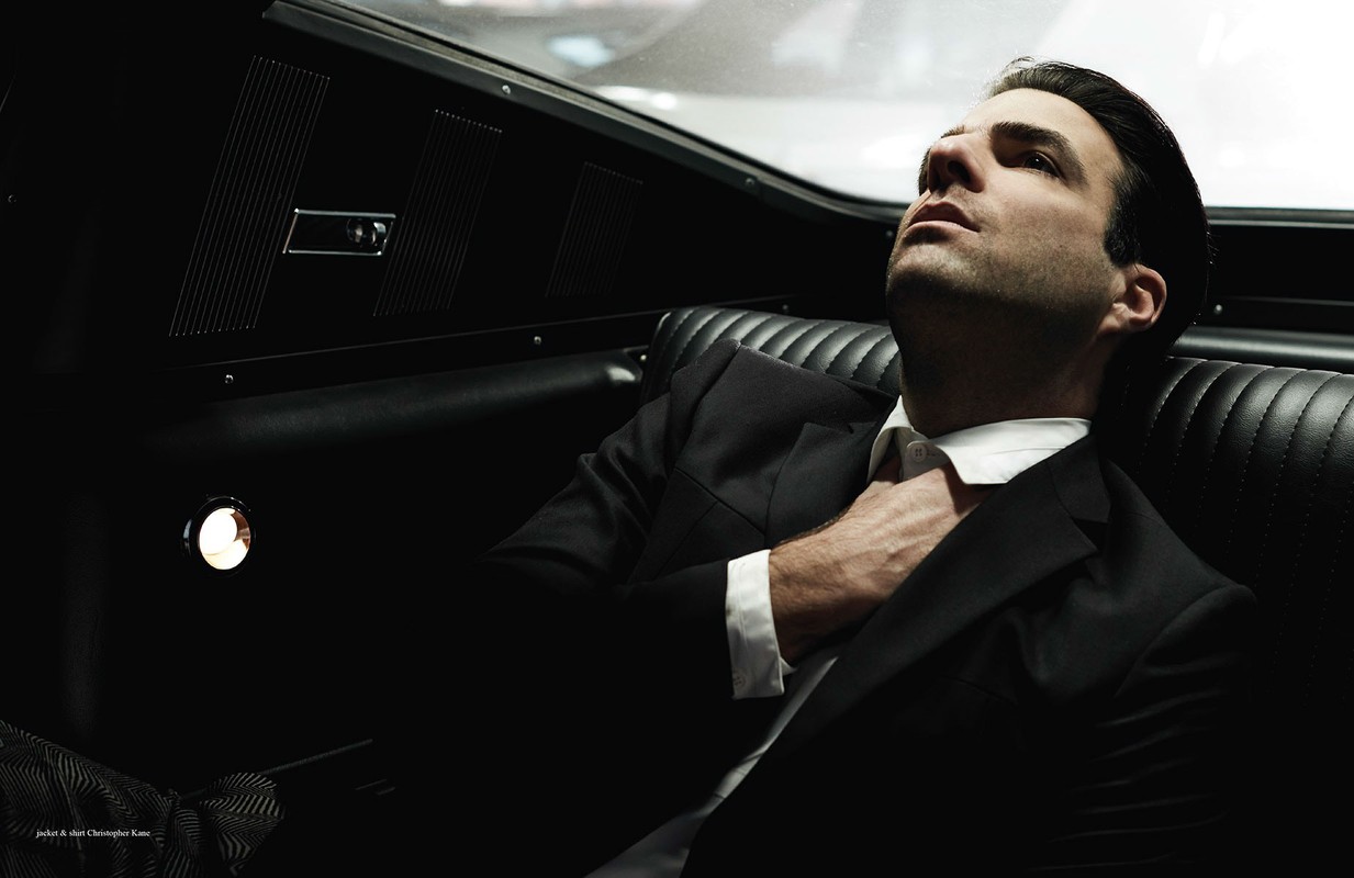 Portrait of a stylish adult male in a black suit reclining in a car interior, looking up with a serious expression in dark, moody lighting.
