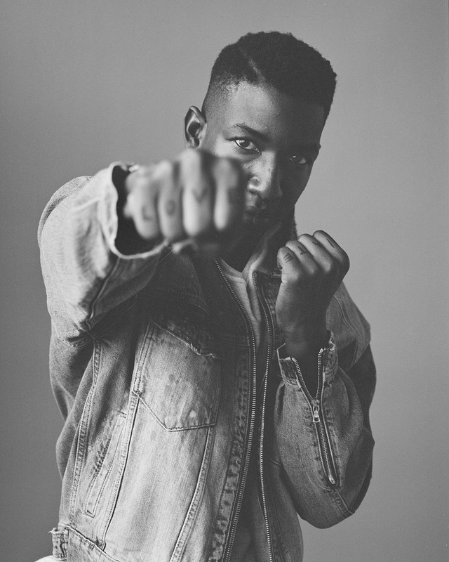 Black and white portrait of a young adult male in a denim jacket, facing forward with a fist raised towards the viewer, indoors.