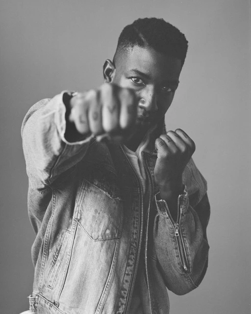 Black and white portrait of a young adult male in a denim jacket, facing forward with a fist raised towards the viewer, indoors.