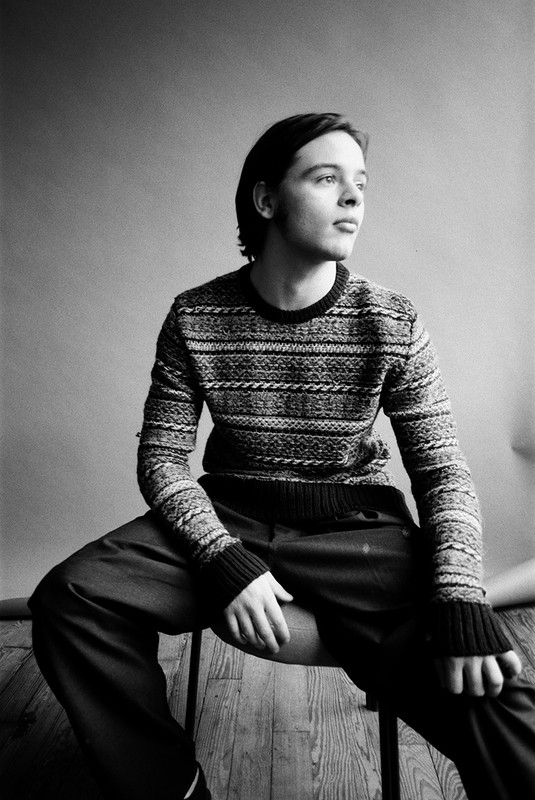 Monochrome portrait of a seated adult male wearing a patterned sweater and dark trousers, gazing off to the side against a plain backdrop.