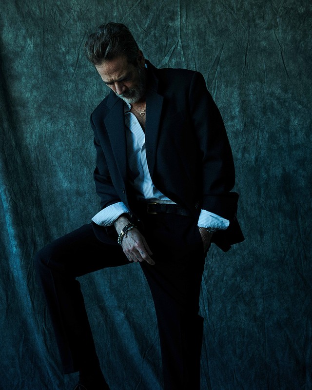 Portrait of a mature adult man posing in a black suit jacket and slacks against a mottled blue-gray studio backdrop with shadow effects.