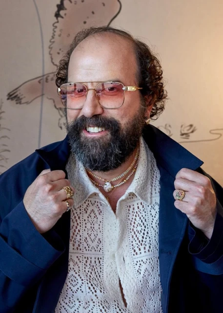Portrait of a bearded adult male with glasses wearing a patterned shirt, rings, and necklaces against a painted backdrop indoors.
