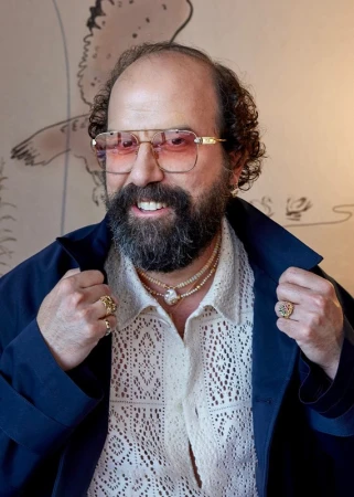 Portrait of a bearded adult male with glasses wearing a patterned shirt, rings, and necklaces against a painted backdrop indoors.