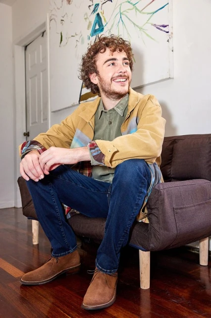 A smiling young man sits on a modern brown couch in casual clothing with artwork visible, set inside of a home with wood floors.