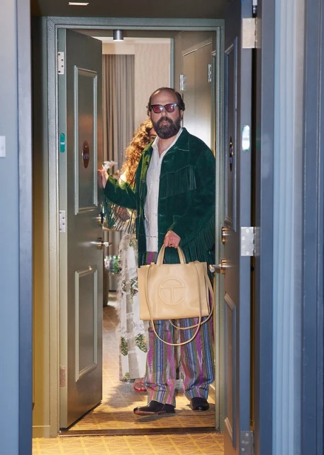 Man in green fringe jacket and patterned pants carrying a tan tote bag stands in a doorway, with a woman visible in the background.