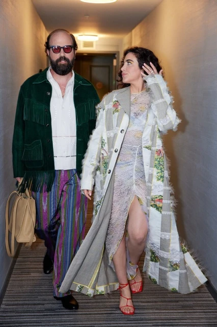 Two people in a hallway, a man with a beard in a fringed jacket and a woman in a patchwork coat, illuminated by overhead lighting.