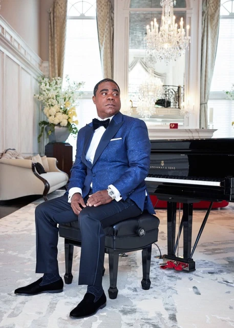 Adult male in a blue suit and bowtie sits on a piano bench in a luxurious interior with a grand piano, chandelier, and white flowers.