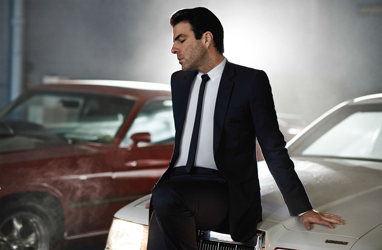 Man in suit leans against vintage car with red car in background, creating a classic and stylish automotive scene with dramatic lighting.