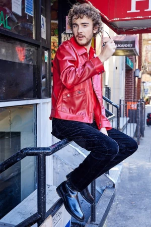 Young man with curly hair in a red jacket and black pants poses on a metal railing in front of a storefront on an urban street.