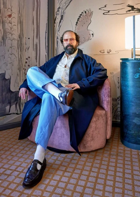 A man with a beard and sunglasses sits in a pink chair wearing a dark coat, light blue pants, and patent-leather shoes, indoors with patterned wallpaper.