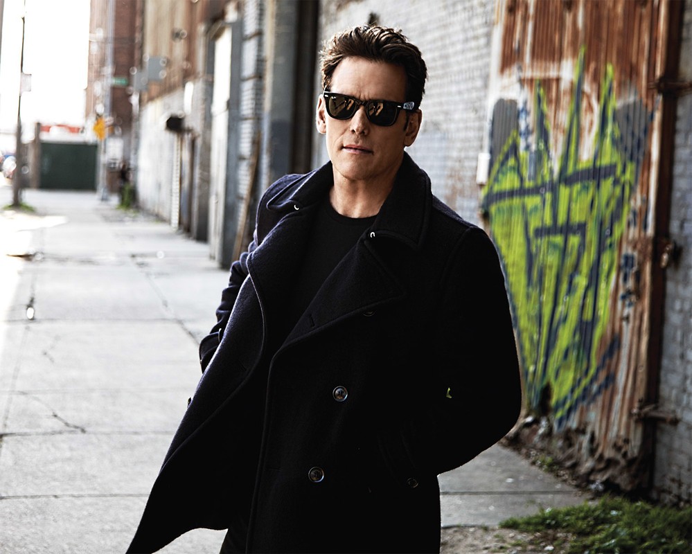 Stylish man in sunglasses and coat poses in an urban alleyway with textured walls, creating a fashionable editorial image.