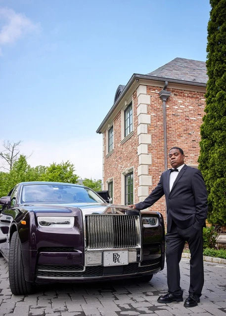 An adult Black male in a suit stands next to a dark purple Rolls Royce in front of a brick mansion on a sunny day.