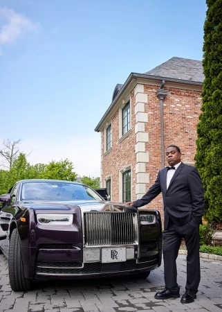 An adult Black male in a suit stands next to a dark purple Rolls Royce in front of a brick mansion on a sunny day.