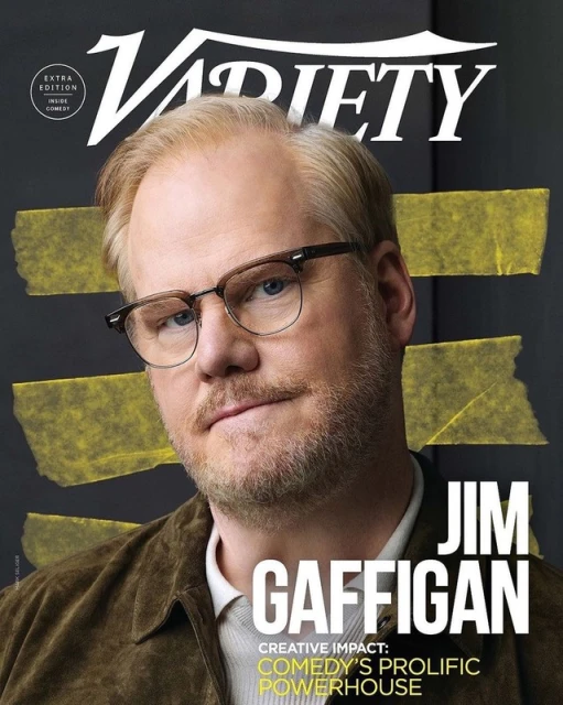 Jim Gaffigan on the cover of Variety Magazine, portrait shot. He's wearing glasses, and a brown jacket. Behind him is a dark background.