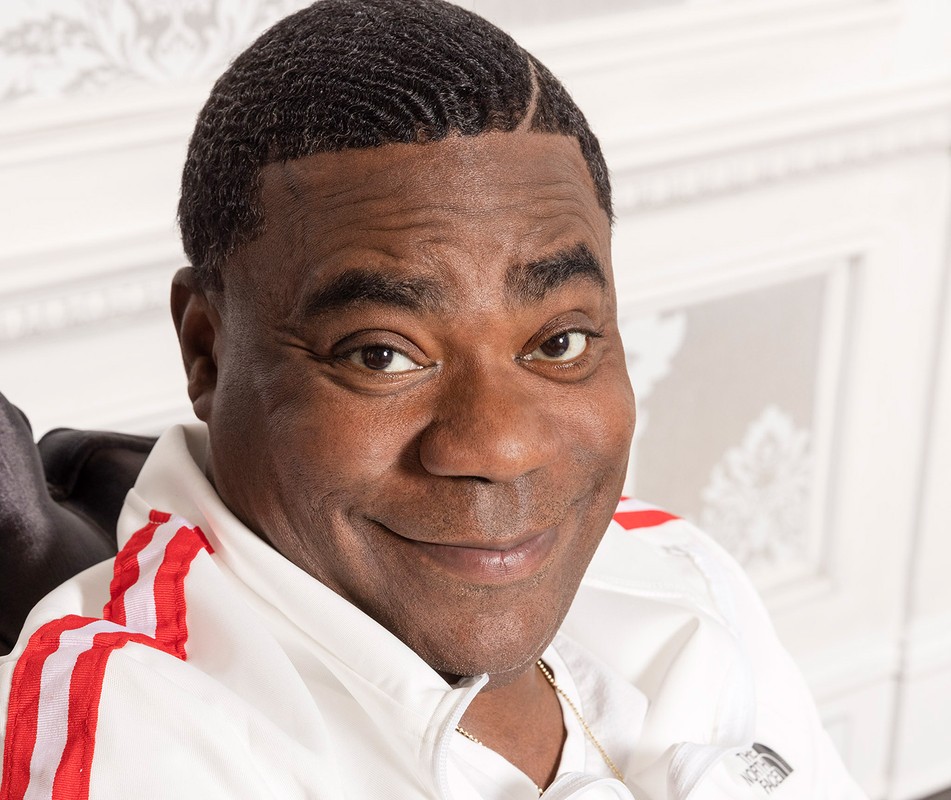 Portrait of an adult man with dark skin smiling while wearing a white jacket with red stripes against an ornate white background.