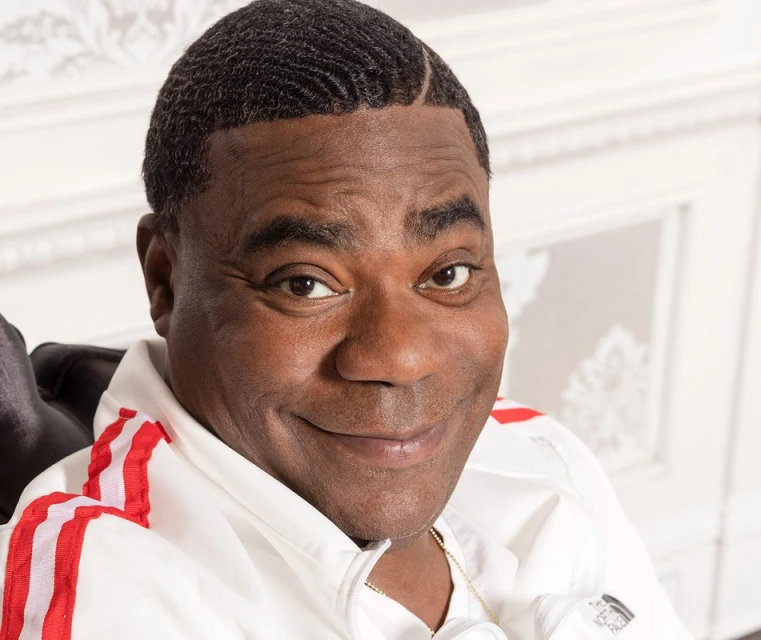 Portrait of an adult man with dark skin smiling while wearing a white jacket with red stripes against an ornate white background.