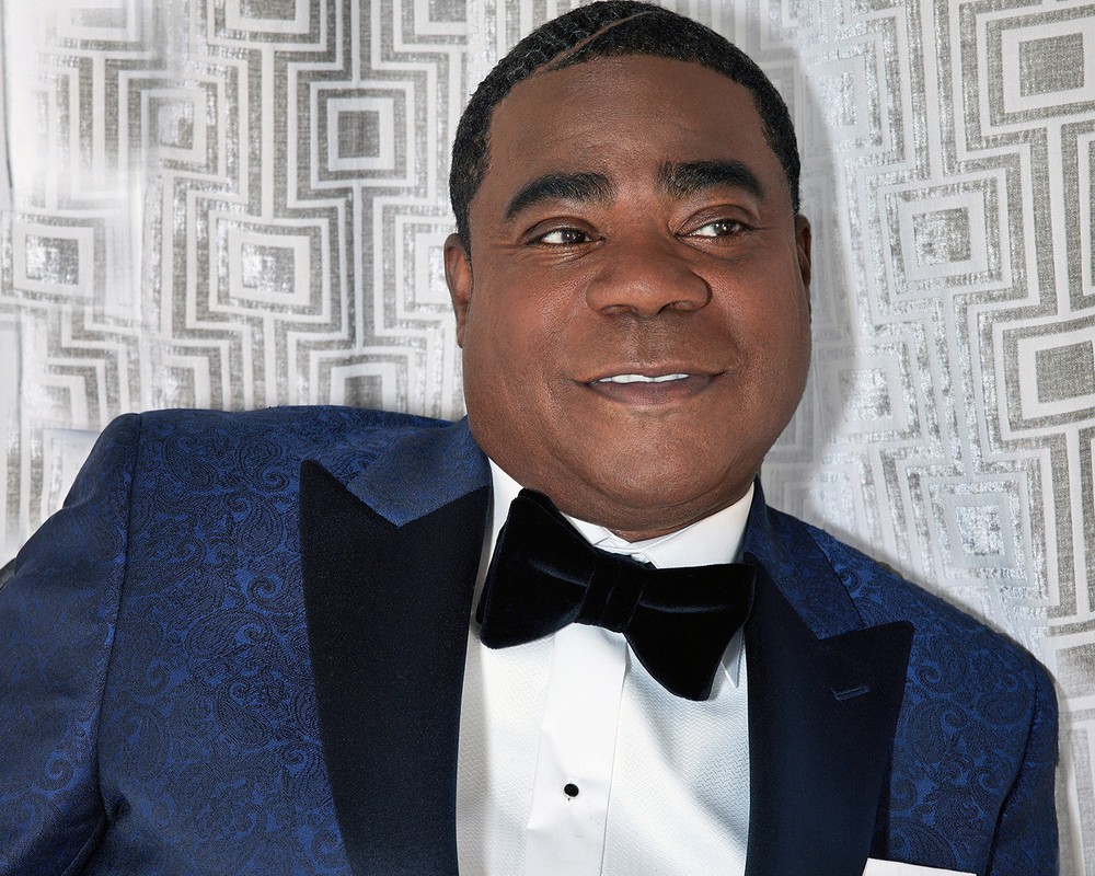 Portrait of adult African-American man in formal blue tuxedo and bow tie posing against grey patterned backdrop in an indoor setting.