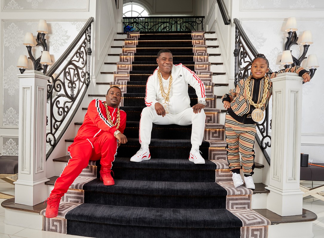 Tracy Morgan, seated on steps with two associates in matching streetwear, gold chains. Indoor setting, likely home interior.