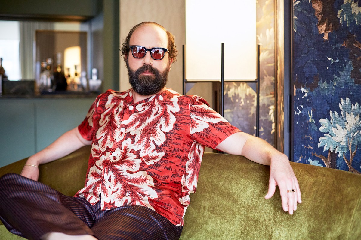 Bearded man in red floral shirt poses on green velvet couch indoors, wearing sunglasses and gold rings, portrait orientation.