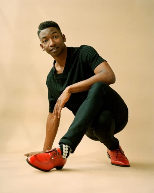Stylish young man in black outfit poses crouching in a studio setting, wearing red shoes. The image captures a modern, fashionable look.