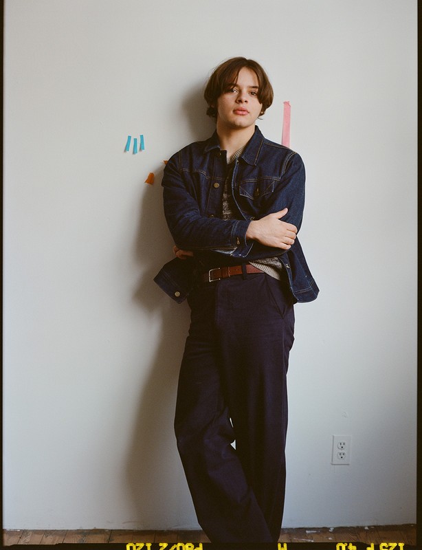 A young adult with shoulder-length brown hair leans against a white wall wearing a dark denim jacket and dark trousers.