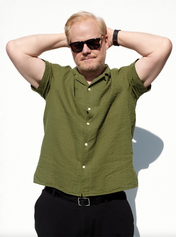 Adult man in a collared green shirt with sunglasses, his hands behind his head, posing against a solid white background in bright sunlight.