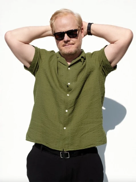 Adult man in a collared green shirt with sunglasses, his hands behind his head, posing against a solid white background in bright sunlight.