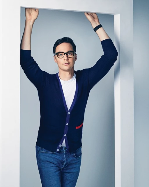 Portrait of Jim Parsons in a doorway wearing glasses, a cardigan, a white T-shirt, and jeans against a grey studio background.