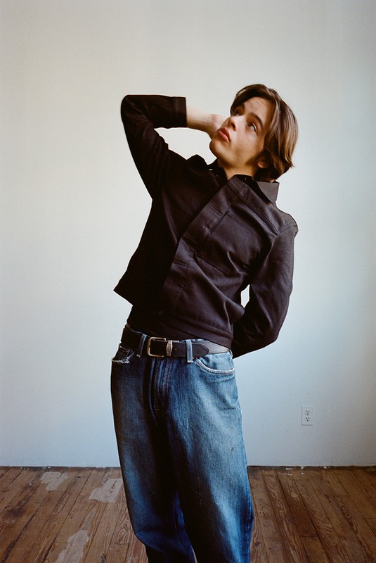 Young adult in a black shirt and blue jeans stands in a light room with a hand behind their head, gazing up at the ceiling.