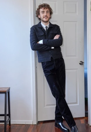 A young man with curly hair wearing a dark textured jacket and tie leaning against a white doorframe.