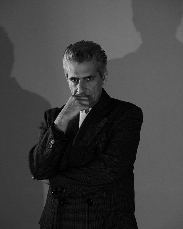 Portrait of a man with graying hair wearing a dark suit, posed thoughtfully with hand to chin, against a neutral backdrop and shadow.