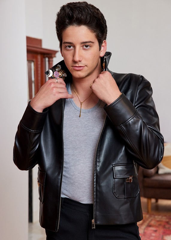 Young man in a black leather jacket poses indoors with a light gray shirt, black pants, gold chain necklace, and multiple statement rings.