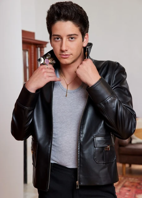 Young man in a black leather jacket poses indoors with a light gray shirt, black pants, gold chain necklace, and multiple statement rings.