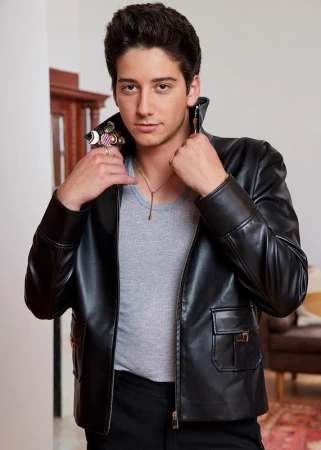 Young man in a black leather jacket poses indoors with a light gray shirt, black pants, gold chain necklace, and multiple statement rings.